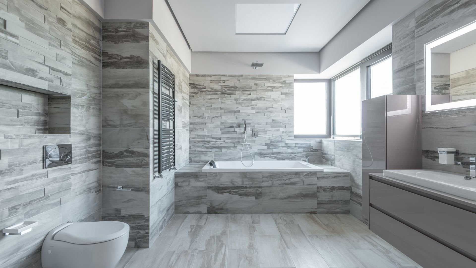 Modern grey bathroom with stone tiles, bathtub, and large windows