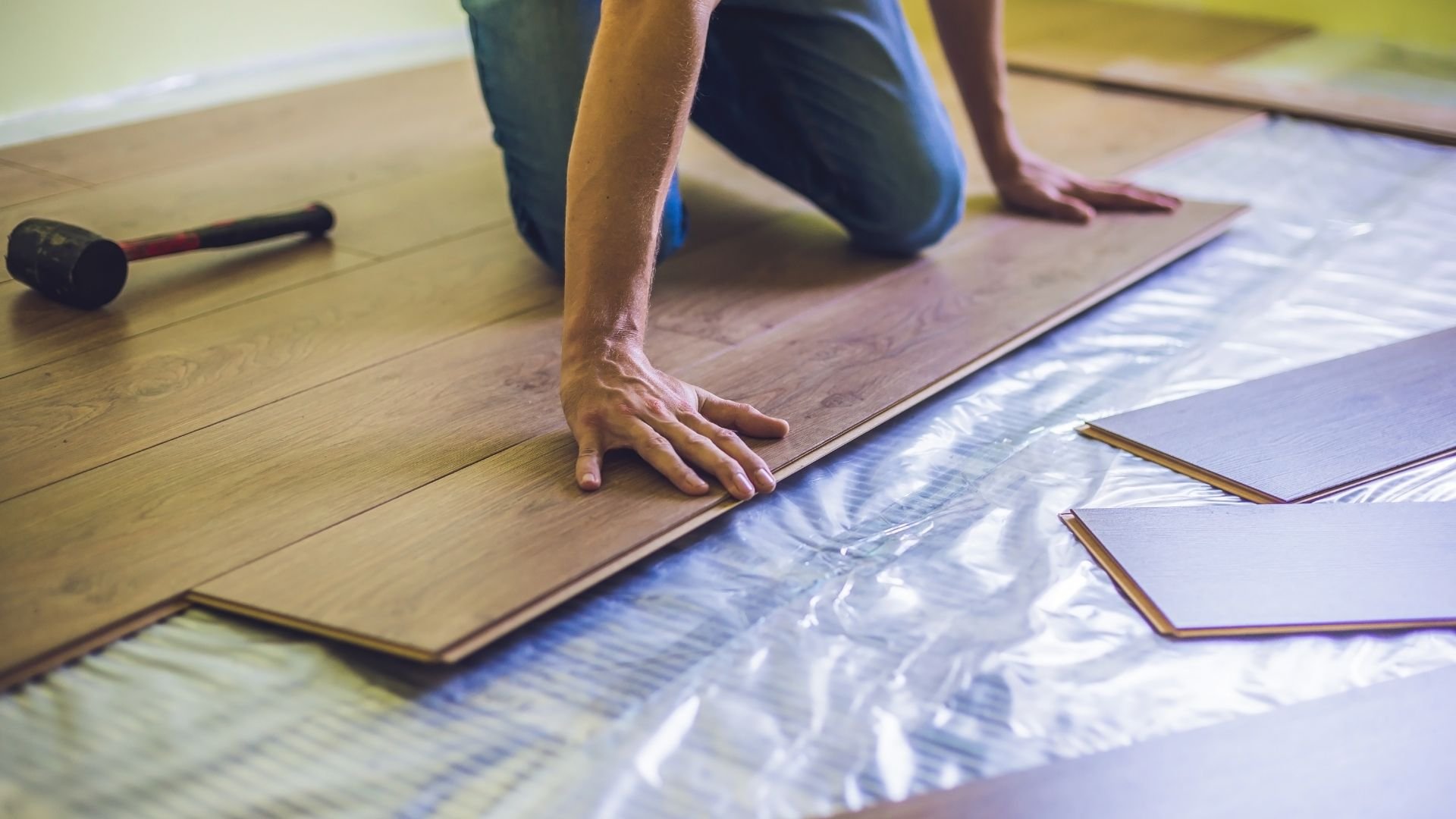 Person installing laminate flooring with rubber mallet and protective underlayment